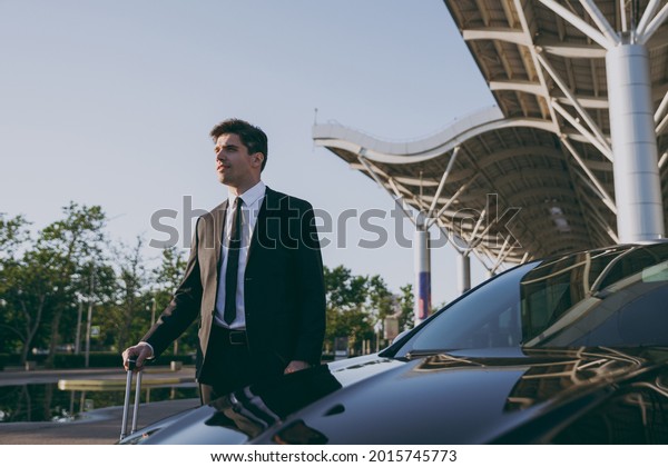 Bottom side view young traveler businessman\
young man in black dinner suit going walk outside at international\
airport terminal with suitcase near car booking taxi Air flight\
business trip concept.