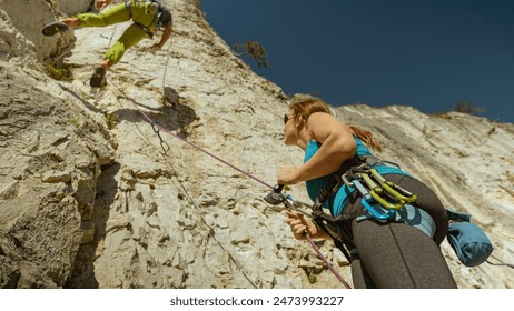 BOTTOM UP, CLOSE UP, DOF: Athletic female belayer helps a fellow rock climber rappel down the rocky cliff. Fit Caucasian woman belays a male rock climber descending from the top of a rocky wall. - Powered by Shutterstock