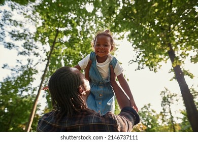 Bottom View Of Adult Black Father Holding And Lifting In Arms Happy Little Daughter In Green Park. Family Relationship And Spending Time Together. Fatherhood And Parenting. Warm Sunny Day