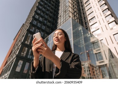 Bottom View Against Background Of High-rise Buildings Young Business Woman With Phone In Her Hands. Long-haired Asian Woman In Transparent Glasses And Stylish Black Suit Is Chatting On Smartphone.