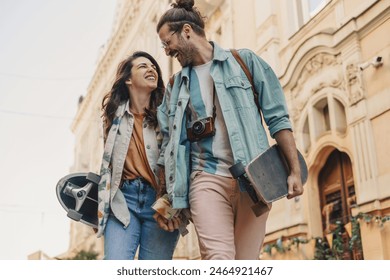 Bottom view of alternative fashionable skateboarder couple holding hands and walking on city street downtown while laughing at each other. Affectionate young hipster couple walking on city street. - Powered by Shutterstock