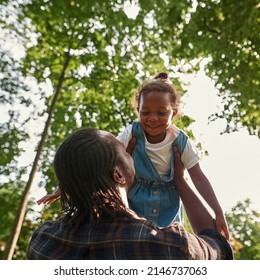 Bottom View Of Black Father Holding And Lifting In Arms Smiling Daughter In Green Park. Man And Little Girl Look At Each Other. Family Relationship And Enjoying Time Together. Fatherhood And Parenting