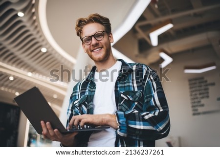 Bottom view of caucasian guy using laptop and looking at camera in office space. Concept of flexible workplace. Idea of freelance and remote work. Young smiling bearded successful man in glasses