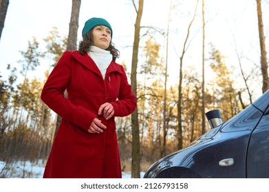 Bottom View Of Confident Dark Haired Woman, Female Traveler Driver Buttoning Up Button On Red Coat While Standing In Front Of Her Car During Coffee Break On The Road, While Travelling In Snowy Nature