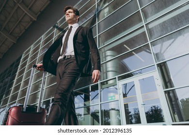 Bottom view confident young traveler businessman man in black dinner suit walk go stand outside at international airport terminal with suitcase valise looking aside. Air flight business trip concept.