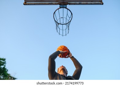 Bottom View Cool Young Sporty Strong Sportsman Man In Sports Clothes Training Shooting Free Throw Play With Ball At Basketball Game Playground Court On Sky Background Outdoor Courtyard Sport Concept.