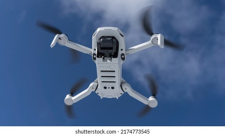 Bottom view of drone with digital camera. White remote controlled quadcopter flying. Close up shot of hovering drone (UAV). Motion blurred propellers. Blue sky with clouds in the background.