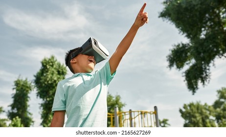 Bottom View Of Excited Boy With Open Mouth Watch Virtual Reality Headset And Point With Finger Up Outdoors. Digital Generation Alpha. Modern Childhood Lifestyle. Gadget Addiction. Sunny Summer Day