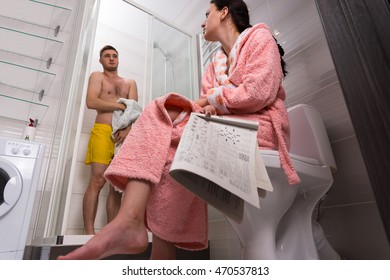 Bottom View Of Female In Bathrobe With Newspaper Sitting On A Toilet And Waiting For Her Boyfriend While He Toweling  In Shower-cabin With Transparent Glass Doors In The Bathroom