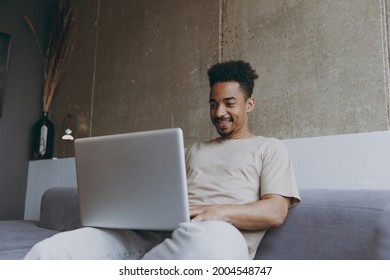 Bottom View Freelancer Young African American Man In Casual Beige T-shirt Sweatpants Sitting On Grey Sofa Indoors Apartment Use Laptop Pc Computer Work Online Programming Resting On Weekends Stay Home