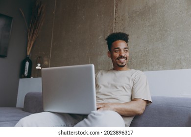 Bottom View Freelancer Young African American Man In Casual Beige T-shirt Sweatpants Sitting On Grey Sofa Indoors Apartment Use Laptop Pc Computer Work Online Look Aside Resting On Weekends Stay Home