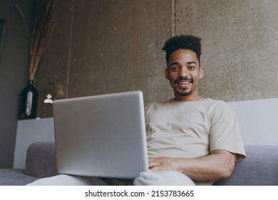 Bottom View Freelancer Young African American Man In Casual Beige T-shirt Sweatpants Sitting On Grey Sofa Indoors Apartment Use Laptop Pc Computer Work Online Look Camera Resting On Weekends Stay Home