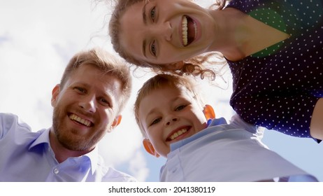 Bottom View Of Happy Mother, Father And Son Look Down At Camera Outdoors. Cheerful Caucasian Parents And Little Boy Smiling At Camera