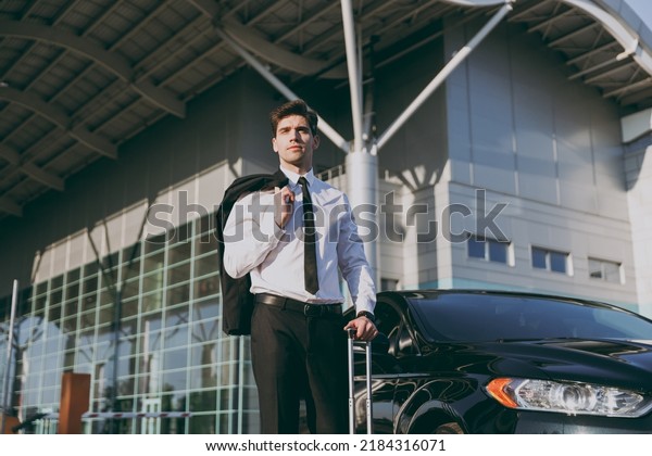 Bottom view pensive young traveler businessman\
young man in black dinner suit going walk outside at international\
airport terminal with suitcase near car booking taxi Air flight\
business trip concept.