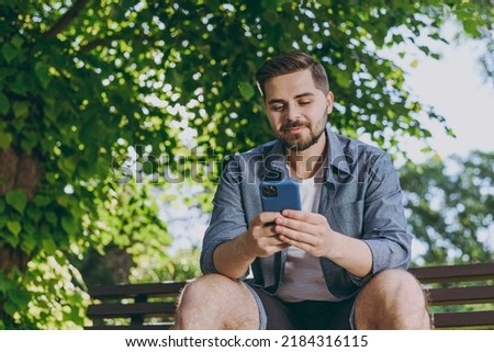 Bottom view smiling happy fun nyoung man in blue shirt sit on bench use mobile cell phone chat online rest relax in spring green city sunshine park outdoors on nature Urban lifestyle leisure concept.