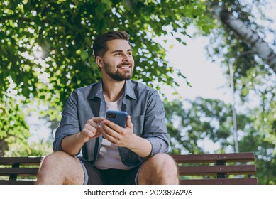 Bottom view smiling minded young man 20s in blue shirt sit on bench use mobile cell phone look aside rest relax in spring green city sunshine park outdoors on nature Urban lifestyle leisure concept.