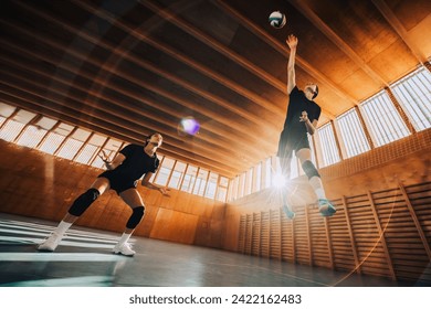 Bottom view of a teenage female professional volleyball players in action hitting a ball on indoor court. Lens flare. Low angle view of a multicultural professional sportswomen hitting a volleyball. - Powered by Shutterstock