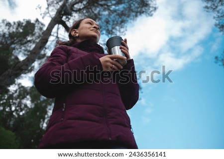 Bottom view. Traveler woman holding a thermos cup with hot drink, tea or coffee, looking away, hiking in the forest. Female adventurer exploring the nature, standing over blue clear sky background