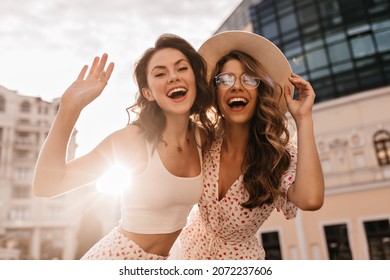 Bottom view of two cheerful beautiful caucasian young women posing for photo in sunbeams. On warm day, brunette and blonde are walking around city in white casual clothes.