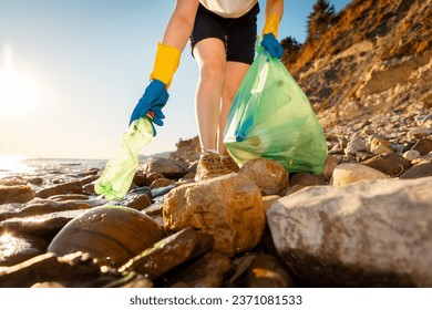 Bottom view of woman volunteer's hand in rubber gloves takes plastic bottle from pebble wild coast. Concept of ocean pollution.