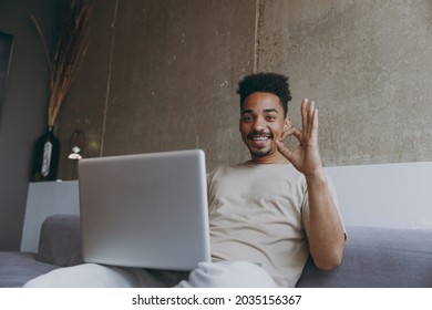 Bottom View Young African American Man In Casual Beige T-shirt Sweatpants Sitting On Grey Sofa Indoors Apartment Use Laptop Pc Computer Work Online Show Ok Okay Gesture Resting On Weekends Stay Home