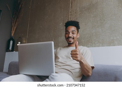 Bottom View Young African American Man In Casual Beige T-shirt Sweatpants Sitting On Grey Sofa Indoors Apartment Use Laptop Pc Computer Work Online Show Thumb Up Gesture Resting On Weekends Stay Home