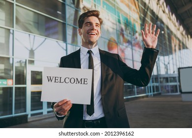 Bottom View Young Cheerful Traveler Businessman Man In Black Suit Stand Outside At International Airport Terminal Hold Card Sign With Welcome Title Text Waving Hand Air Flight Business Trip Concept.