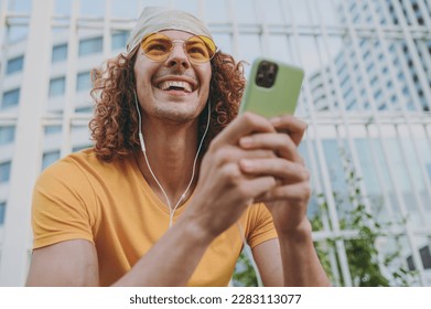 Bottom view young cool man 20s in yellow t-shirt bandana talk speak on mobile cell phone listen to music in headphones sitting rest relax in city outdoors on open air. Urban lifestyle leisure concept - Shutterstock ID 2283113077