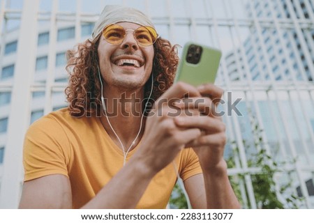 Bottom view young cool man 20s in yellow t-shirt bandana talk speak on mobile cell phone listen to music in headphones sitting rest relax in city outdoors on open air. Urban lifestyle leisure concept