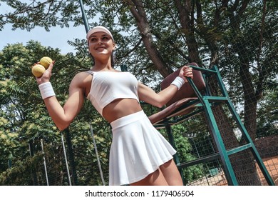 Bottom View Of Young Female Model Wearing Fashionable Tennis Sportwear, Posing On Tennis Judge Umpire Chair With Tennis Balls, Summer Sunny Day In Sport Club