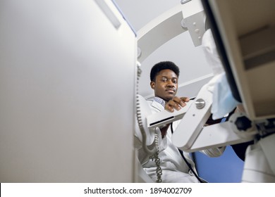 Bottom view of young handsome high-skilled African doctor in white coat, working with modern lithotripter equipment for non-invasive extracorporeal shock wave lithotripsy