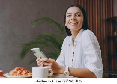 Bottom View Young Smiling Minded Happy Cheerful Fun Latin Woman 30s Wear White Shirt Use Mobile Cell Phone Sit Alone At Table In Coffee Shop Cafe Restaurant Indoors. Freelance Office Business Concept