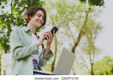 Bottom view young smiling student freelancer woman 20s in green jacket jeans sit on bench in spring park outdoors rest use laptop pc computer hold mobile cell phone. People urban lifestyle concept.