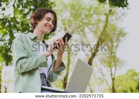 Bottom view young smiling student freelancer woman 20s in green jacket jeans sit on bench in spring park outdoors rest use laptop pc computer hold mobile cell phone. People urban lifestyle concept.