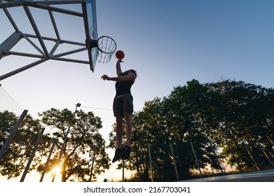 Bottom View Young Strong Sportsman Man In Sports Clothes Training Shooting Free Throw Fly High Jump Scoring Basket Play With Ball At Basketball Game Playground Court Outdoor Courtyard Sport Concept.