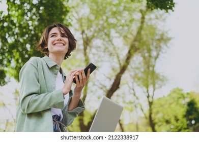 Bottom-View junge Studentin Freelancer Frau in grüner Jacke Jeans sitzen auf Bank im Frühlingspark im Freien Ruhe Laptop PC-Gespräch über Handy Blick zur Seite. Stadtleben