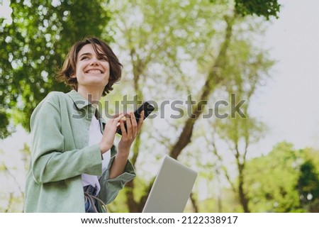 Bottom view young student freelancer woman in green jacket jeans sit on bench in spring park outdoors rest use laptop pc computer talk by mobile cell phone look aside. People urban lifestyle concept