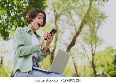 Bottom View Young Surprised Student Freelancer Woman In Green Jacket Jeans Sit On Bench In Spring Park Outdoors Rest Use Laptop Pc Computer Mobile Cell Phone Look Aside. People Urban Lifestyle Concept