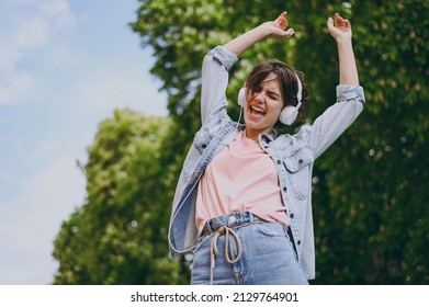 Bottom view young woman in jeans clothes headphones listen to music walk dance in green park dance with raised up hands outdoors with green blur trees on background. People urban lifestyle concept. - Shutterstock ID 2129764901