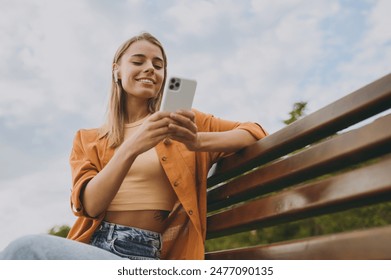 Bottom view young woman wear orange shirt casual clothes use mobile cell phone listen music in earphones sit on bench walk rest relax in spring city park outdoors on nature. Urban lifestyle concept - Powered by Shutterstock