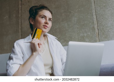 Bottom View Young Woman In White Clothes Sit On Grey Sofa Indoors Apartment Use Mobile Cell Phone Credit Bank Card Shopping Online Order Delivery Booking Tour Rest On Weekend Leisure Stay Home Concept