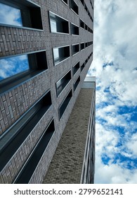 A bottom-up perspective looking at the facade of a modern multi-story brick residential or office building against a blue sky. The image is focused on the corner of a tall skyscraper or apartment complex. The facade is clad in brown and gray textured clinker bricks arranged in neat geometric patterns. Long vertical columns of rectangular windows with deep dark metal frames run the length of the building. The glass panes of the windows reflect the blue sky and soft white clouds. The upper right of the frame is dominated by a vast, dramatic sky filled with scattered high cumulus clouds. The composition with diagonal and vertical lines creates a powerful sense of height, scale and modern urban design.