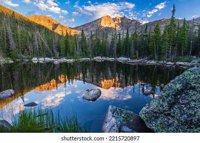 Boulders surround the calm water of Chipmunk Lake reflecting Ypsilon Mountain and Mount Chiquita lit by morning light in Rocky Mountain National Park, Colorado.