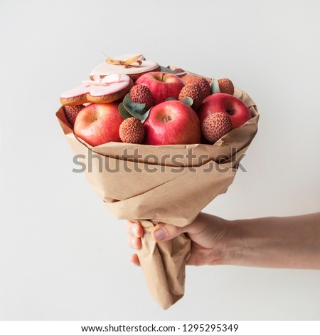 Bouquet of apples and lychee with  
gingerbread cookies in shape of heart on a white background. Hand holding a beautiful fruit bouquet.  Gift for lover. 