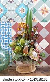 Bouquet of beautiful bush flowers including king protea, protea, banksia, kangaroo paw, and eucalyptus leaves against a colorful tiled wall with a part green glass vase visible next to the arrangement