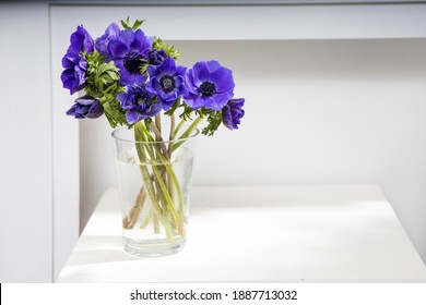 bouquet of the blue anemones in a glass vase on a white table near the pale gray wall. Vertical frame. Copy space.