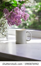 A bouquet of flowers of lilacs on the window and coffee in a white cup in the rays of sunlight. A bouquet of lilacs and a cup of coffee or tea.  - Shutterstock ID 1985876942