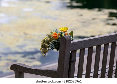 A Bouquet Of Flowers On A Park Bench Next To Water. In Memory, Remembrance.