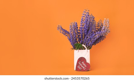 Bouquet of fresh lupine flowers in white vase shaped like bag and wooden red heart with white inscription happy Father's day on orange background - Powered by Shutterstock