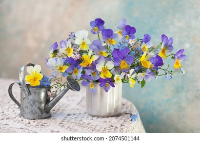 A bouquet of pansies and forget-me-not flowers in a vase and a watering can on a table with a tablecloth against the background of a colored wall. Romantic postcard, blur, selective focus.